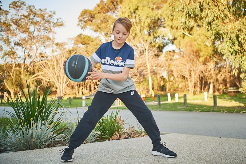 A young boy bounces a basketball in his driveway. The sun is setting on the trees in the background.