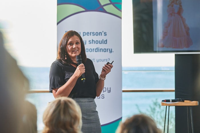 Neryl Joyce stands on the stage and addresses the audience. She is standing in front of a banner and tv screen with Therapy Focus branding. Behind her is the ocean. 