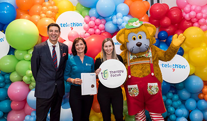Two therapists stand with Basil Zempilas and Fat Cat in front of a rainbow balloon wall.