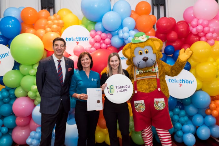 Two therapists stand with Basil Zempilas and Fat Cat in front of a rainbow balloon wall. One of the therapists is holding a certificate and the other is holding a Therapy Focus branded sign.