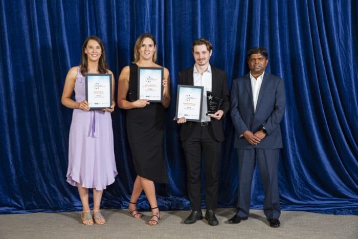 Katy, Emily, Hamish and Kanishka stand in front of a blue background and smile. 