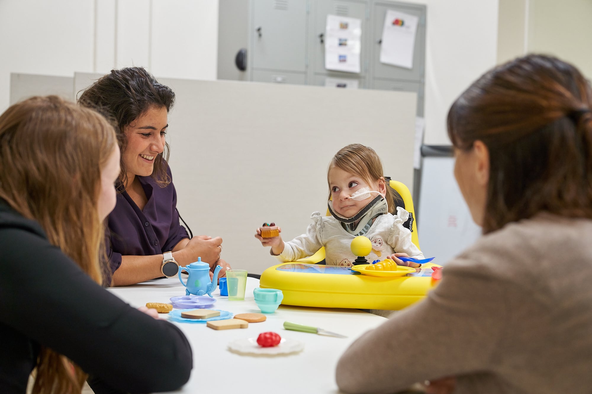 3 adults support a young child as she engages in play. The young child sits in a yellow Explorer Mini assistive technology device. She has light brown hair and uses a feeding tube and neck brace.